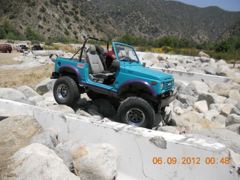 1986 Ford Bronco - A blue Suzuki Samurai tackles a challenging off-road course filled with large rocks and concrete barriers.
