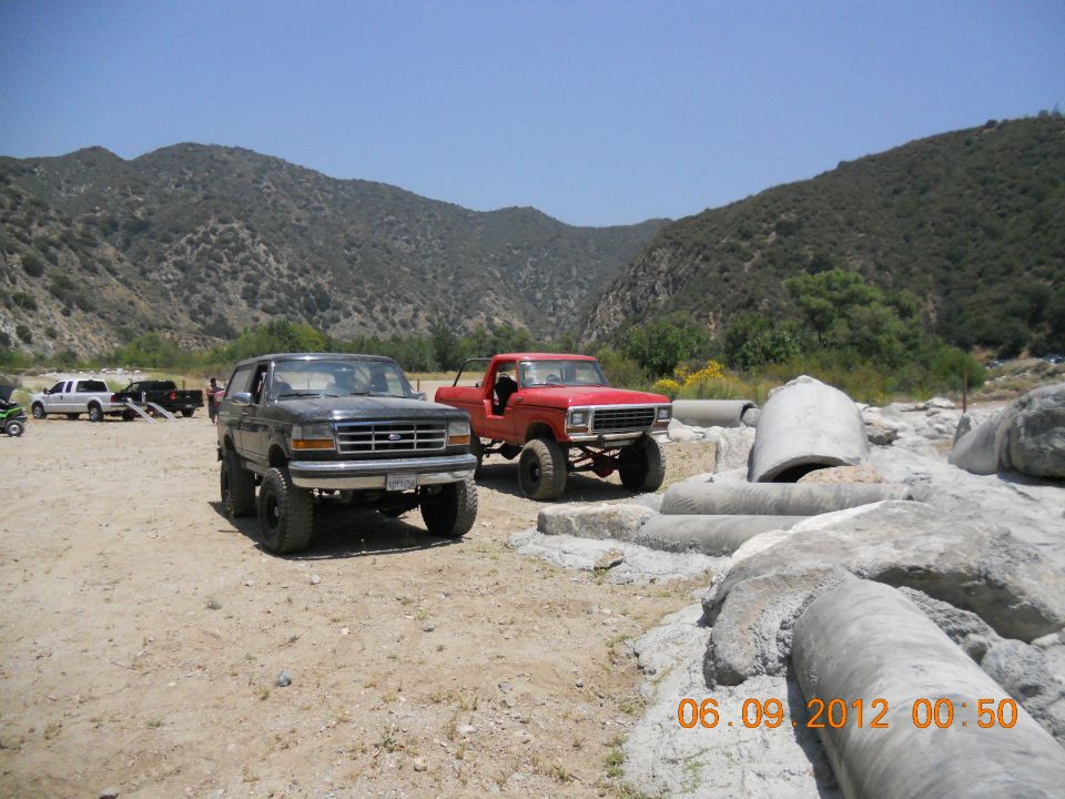 Lifted black Bronco and red pickup truck parked in a dry riverbed with mountains behind them