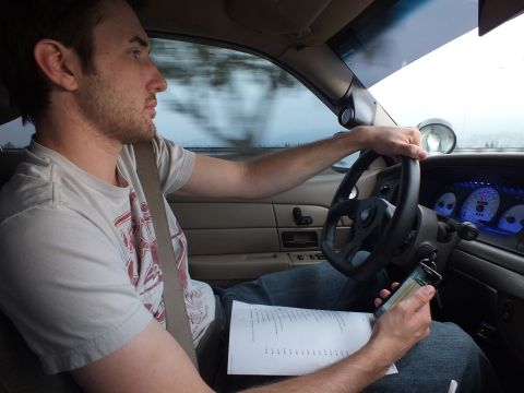2003 Ford Crown Victoria - Driver in a gray tee steers while holding documents, blue-lit gauges glowing at right.