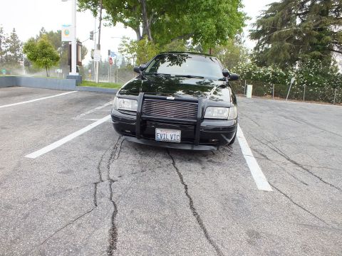 2003 Ford Crown Victoria - Front of a blacked-out Crown Victoria fitted with a police-style push bumper, parked on cracked asphalt.