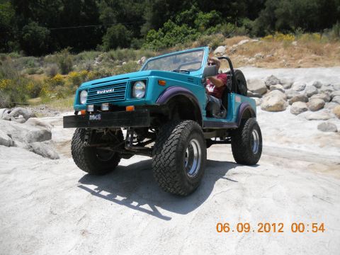 1986 Ford Bronco - Suzuki Samurai off-roading on a sunny day with rocks and dry brush in the background.