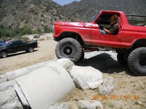 1986 Ford Bronco - A red off-road truck is shown navigating a rocky terrain, with another pickup truck parked in the background.