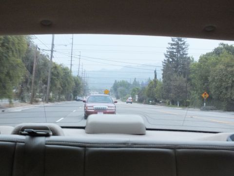 2003 Ford Crown Victoria - Maroon Lincoln sedan traveling ahead on a wide road framed by a car's rear glass and beige headrest.