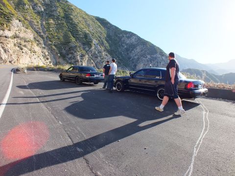 2003 Ford Crown Victoria - Two black sedans and a few people gathered at a sunlit mountain pullout with steep ridges behind.