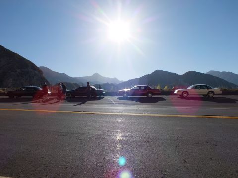2003 Ford Crown Victoria - Several cars line a roadside pullout, backlit by a low sun over a jagged mountain skyline.
