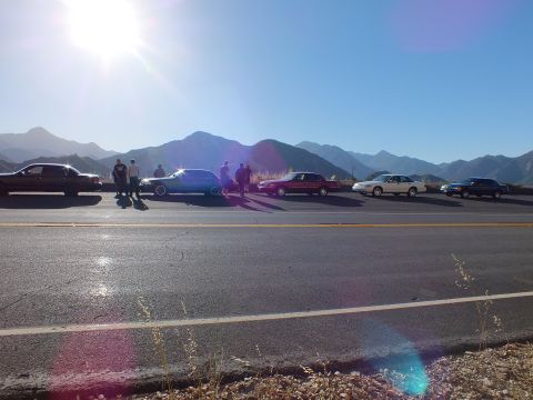 2003 Ford Crown Victoria - string of sedans and onlookers spread along a roadside pullout under bright sun and rugged peaks.