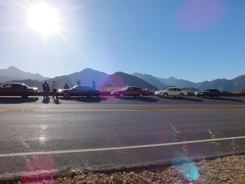 2003 Ford Crown Victoria - Backlit lineup of full-size sedans on a roadside with people and layered mountains behind.