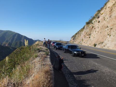 2003 Ford Crown Victoria - Lineup of dark full-size sedans on a cliffside road; people stand at the overlook as one crouches to photograph the cars.