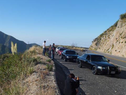 2003 Ford Crown Victoria - Dark Crown Victoria-style sedan at a roadside overlook; a person kneels photographing the parked lineup against distant peaks.