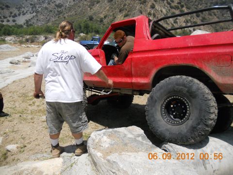 1986 Ford Bronco - A red off-road SUV is seen navigating a rocky terrain with a person standing nearby. Another vehicle is visible in the background.