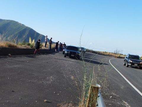 2003 Ford Crown Victoria - Group gathered beside a dark sedan on a freshly paved overlook; an SUV rounds the curve on the highway beyond.