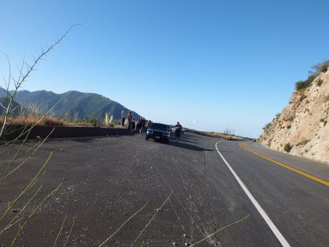 2003 Ford Crown Victoria - Group stands near a parked SUV on a broad turnout; the road curves along the ridge under bright sky.