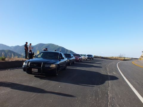 2003 Ford Crown Victoria - Dark sedan with brush guard leads a row of parked cars at a paved overlook above a hazy valley.