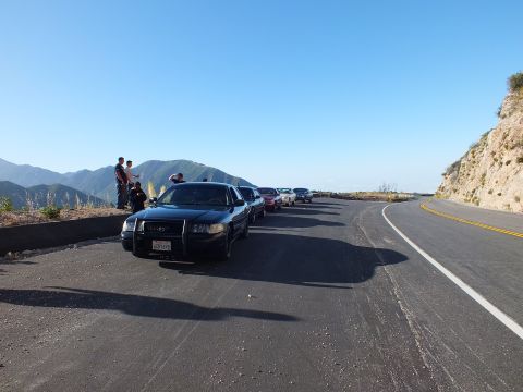 2003 Ford Crown Victoria - Dark sedan at the front of a parked lineup on a wide turnout; the highway bends along the sunlit ridge.