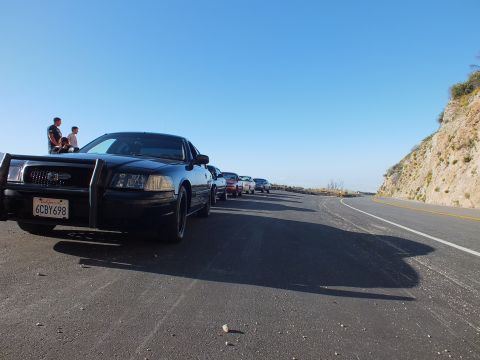 2003 Ford Crown Victoria - Low front-quarter shot of a dark sedan with brush guard; two people sit on the hood at a ridge turnout.
