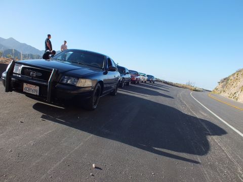 2003 Ford Crown Victoria - Dark sedan with brush guard at the front of a parked row; two people perch on the hood at a mountain overlook.
