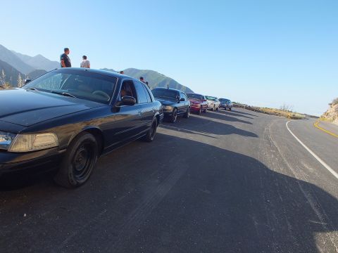 2003 Ford Crown Victoria - Low-angle view of a dark sedan heading a parked lineup; people stand at the ridge as the road curves away.