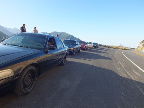 2003 Ford Crown Victoria - Side-front view of a dark sedan leading parked cars at a mountain overlook; people gather near the edge.