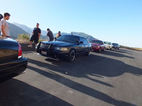 2003 Ford Crown Victoria - Dark sedan with brush guard in a paved lot; a red sedan and more cars line the ridge as onlookers gather.