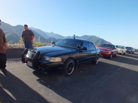 2003 Ford Crown Victoria - Front-quarter view of a blacked-out sedan with brush guard; a red sedan and others sit along the sunlit ridge.
