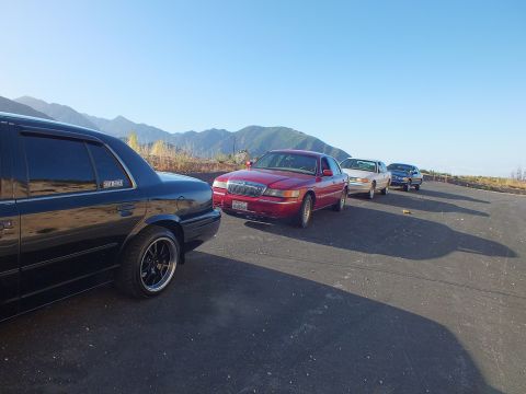 2003 Ford Crown Victoria - Black sedan in foreground with a red Grand Marquis and lighter cars lined up along a ridge turnout.