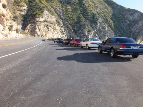 2003 Ford Crown Victoria - Cars lined along a curving mountain road, people standing between them, hills rising behind