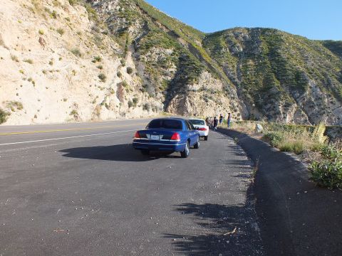 2003 Ford Crown Victoria - Two sedans on a broad paved shoulder, several people standing by the guardrail under green hills