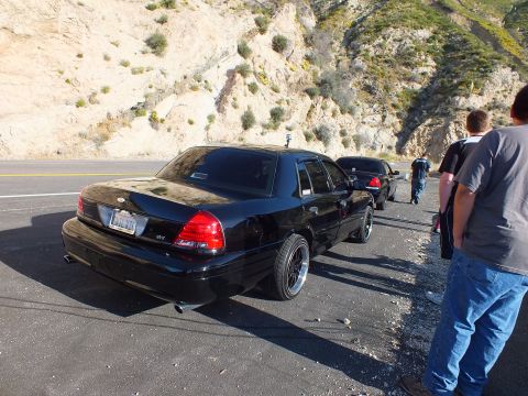 2003 Ford Crown Victoria - Lowered black sedan with dual exhaust and police-style spotlight, group of men standing at right
