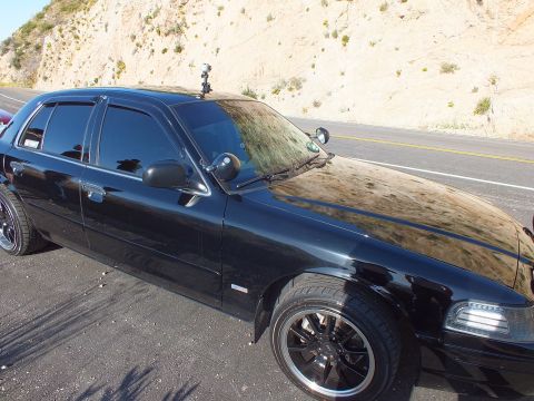 2003 Ford Crown Victoria - Glossy black four-door close up showing spotlight, side mirror camera and dark alloy wheels