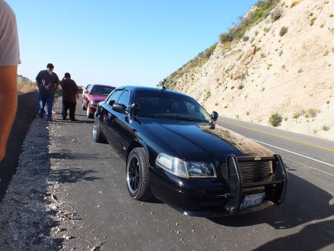 2003 Ford Crown Victoria - Lowered black sedan with push bar and spotlight, people and a red car behind on the shoulder