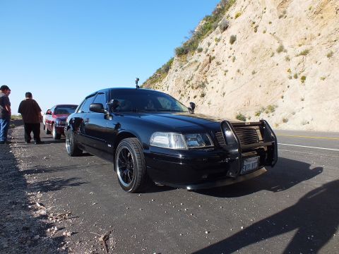 2003 Ford Crown Victoria - Black sedan with brush guard and spotlight, red sedan and onlookers at left under rocky bluff
