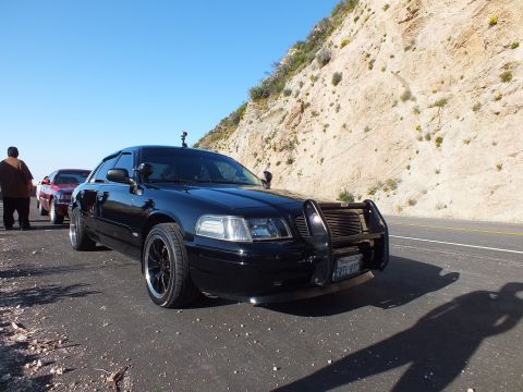 2003 Ford Crown Victoria - Front three-quarter of a lowered black sedan with brush guard, hillside and shadows behind