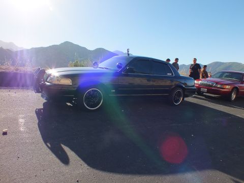 2003 Ford Crown Victoria - Lowered black four-door silhouetted against sun glare, red sedan and people to the right