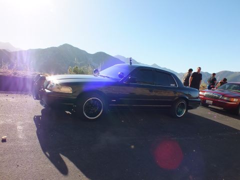 2003 Ford Crown Victoria - Black four-door catching sunburst flare, red sedan and onlookers beside mountain backdrop