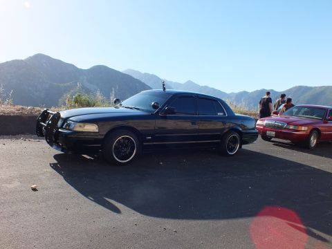 2003 Ford Crown Victoria - Black four-door with brush guard parked by a red sedan, people at right and peaks behind