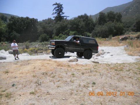 1986 Ford Bronco - A black Ford Bronco is parked on a dirt path in a wooded, mountainous area. A person stands to the left of the vehicle.