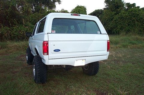 1995 Ford Bronco - Rear view of a white Ford Bronco full-size SUV with a hardtop, lifted suspension and chunky off-road tires, parked on grass in front of den…