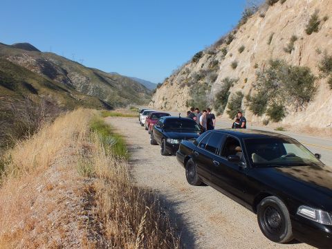 2003 Ford Crown Victoria - Dark sedans queued on a gravel turnout, onlookers chatting beside the road under blue sky