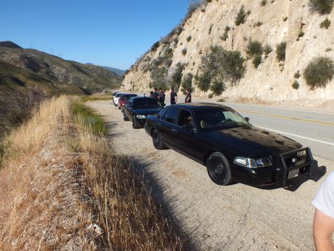 2003 Ford Crown Victoria - Dark full-size sedan with push bar parked along a mountain highway, group gathered beside the road