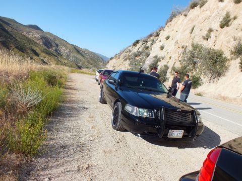 2003 Ford Crown Victoria - Front three-quarter of a blacked-out Ford sedan on a dirt pullout, more vehicles lined up behind