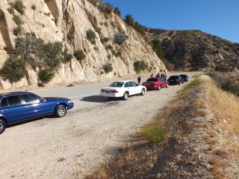 2003 Ford Crown Victoria - Row of sedans and a small truck on a dirt shoulder, people gathered near the rock cliff