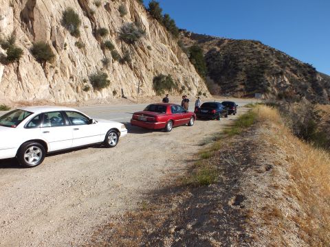 2003 Ford Crown Victoria - White sedan and maroon sedan on a dirt turnout, dark cars and people beyond against rock face