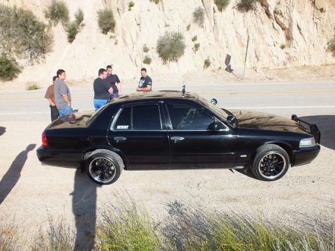 2003 Ford Crown Victoria - Side profile of a lowered black sedan on aftermarket wheels, group of men talking behind it