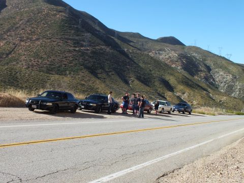 2003 Ford Crown Victoria - Two dark sedans and a group of people on a roadside shoulder, power lines crossing the ridge
