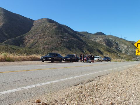 2003 Ford Crown Victoria - Dark sedans parked along a mountain road with people standing between them near a 30 MPH sign