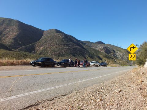 2003 Ford Crown Victoria - Lineup of dark and red cars with people on a mountain road shoulder near a curve warning sign