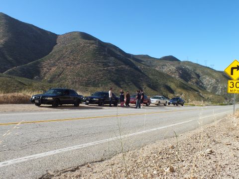 2003 Ford Crown Victoria - Dark sedans and a small red car parked along a mountain road, group of people standing nearby
