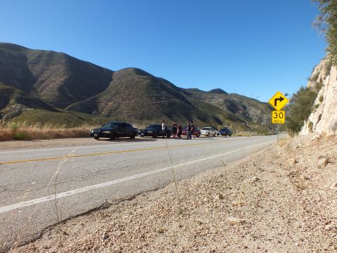 2003 Ford Crown Victoria - Mountain highway with a cluster of parked cars and people on the opposite shoulder near a curve sign