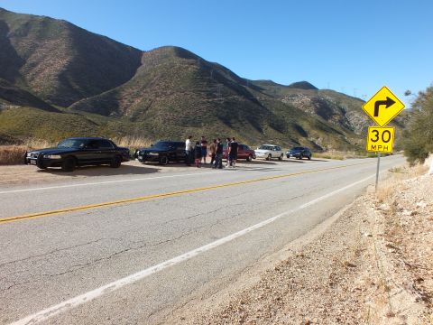 2003 Ford Crown Victoria - Dark sedans parked along a mountain road with people gathered between them near a curve sign
