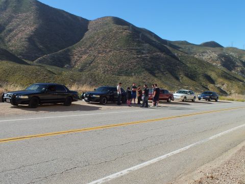 2003 Ford Crown Victoria - Two black sedans and a crowd of people on a mountain road turnout, other cars parked beyond